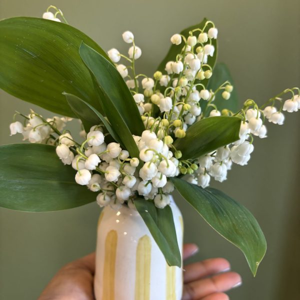 Bouquet de muguet du 1er mai dans un petit vase artisanal rayé jaune et blanc tenu dans une main - Flormandise Vernioz